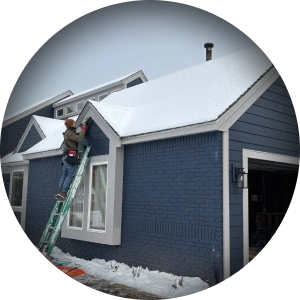 Crew member on a ladder at the eave of a snow-covered roof on a navy house